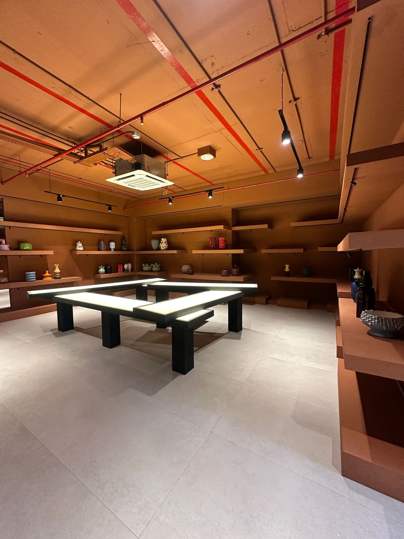 Art and decor store with rust-toned walls, floating timber shelves and backlit display tables under an exposed red-painted ceiling