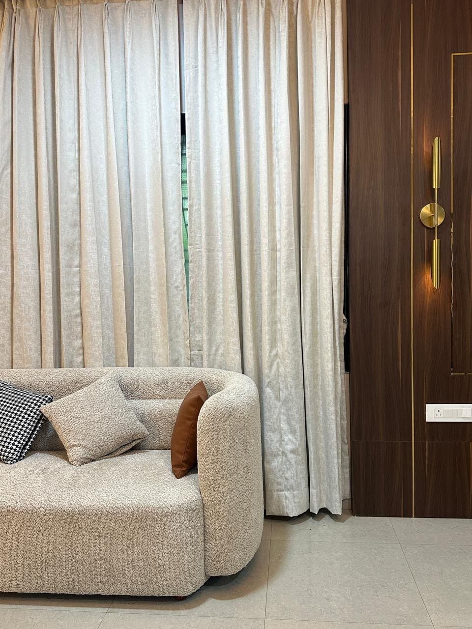 Curved boucle sofa against sheer curtains, beside a walnut panel with a brass sconce