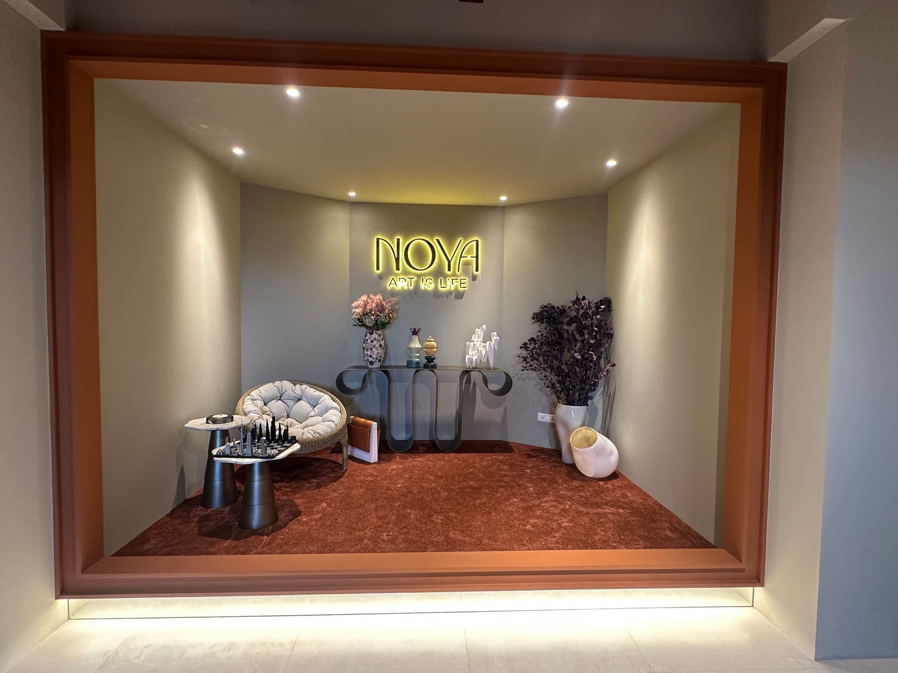 Window vitrine with a neon sign, a papasan chair, a chess set and dried flowers on a tan carpet
